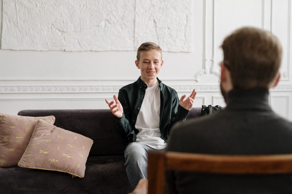 A younger white male sites on a couch talking to an counsellor