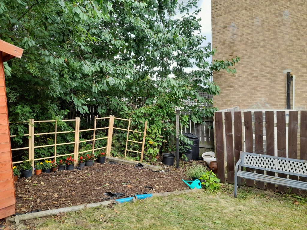 A tidied garden space, with trimmed foliage, new fences and a bench, and a new bed ready for planting.